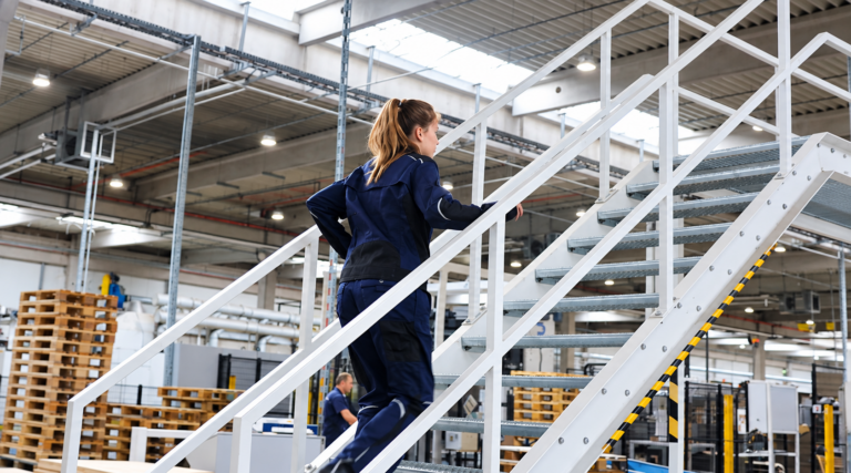 Eine Frau in dunkelblauer Arbeitskleidung steigt eine weiße Metalltreppe in einer modernen Industriehalle hinauf. Im Hintergrund sind gestapelte Holzpaletten, Maschinen und hohe Decken mit sichtbaren Leitungen und Beleuchtung zu sehen. Die Halle wirkt geräumig und gut ausgeleuchtet.