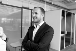 Ein Mann im Anzug steht mit verschränkten Armen in einem modernen Büro. Er lächelt und blickt leicht zur Seite. Im Hintergrund sind Glastrennwände, ein Stuhl und ein Teil eines weiteren Arbeitsplatzes zu sehen. Die Aufnahme ist in schwarz-weiß gehalten.