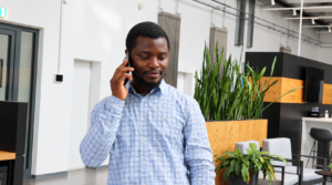 Ein Mann mit kurzem Bart und dunklem Haar steht in einem modernen Büro und führt ein Telefongespräch. Er trägt ein hellblau kariertes Hemd und hält das Smartphone am Ohr. Im Hintergrund sind hohe grüne Pflanzen, Holzelemente und helle Wände zu sehen. Die Einrichtung wirkt offen und lichtdurchflutet.