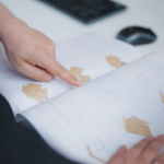 Hand zeigt auf geöffnete Broschüre mit Verpackungsskizzen auf Schreibtisch mit Computermaus im Hintergrund