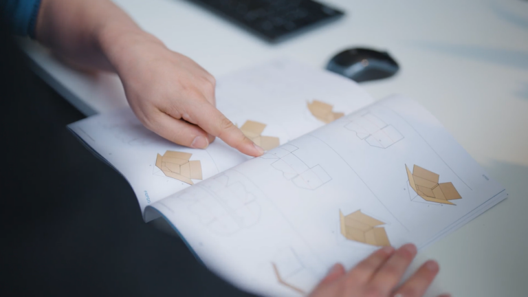 Hand zeigt auf geöffnete Broschüre mit Verpackungsskizzen auf Schreibtisch mit Computermaus im Hintergrund