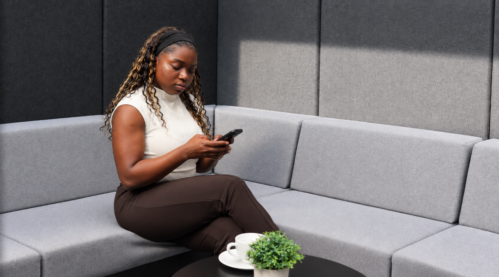 Eine Frau mit langen, lockigen Haaren sitzt auf einem hellgrauen, gepolsterten Sofa in einer modernen Lounge. Sie trägt ein ärmelloses, helles Oberteil und dunkle Hose. Ihr Blick ist auf ein Smartphone gerichtet, das sie mit beiden Händen hält. Vor ihr steht ein runder, schwarzer Tisch mit einer weißen Tasse, einer Untertasse und einer kleinen, grünen Topfpflanze. Die Wände im Hintergrund sind mit grauen, gepolsterten Paneelen verkleidet.