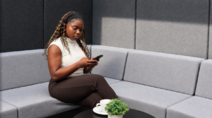 Eine Frau mit langen, lockigen Haaren sitzt auf einem hellgrauen, gepolsterten Sofa in einer modernen Lounge. Sie trägt ein ärmelloses, helles Oberteil und dunkle Hose. Ihr Blick ist auf ein Smartphone gerichtet, das sie mit beiden Händen hält. Vor ihr steht ein runder, schwarzer Tisch mit einer weißen Tasse, einer Untertasse und einer kleinen, grünen Topfpflanze. Die Wände im Hintergrund sind mit grauen, gepolsterten Paneelen verkleidet.