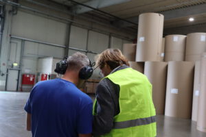 Zwei Mitarbeitende stehen in einer Produktionshalle eines Wellpappenherstellers und tauschen sich aus. Eine Person trägt ein blaues T-Shirt und einen Gehörschutz, die andere eine gelbe Warnweste mit reflektierenden Streifen. Im Hintergrund sind mehrere große, stehende Papierrollen zu sehen, die als Rohmaterial für die Herstellung von Wellpappe dienen.